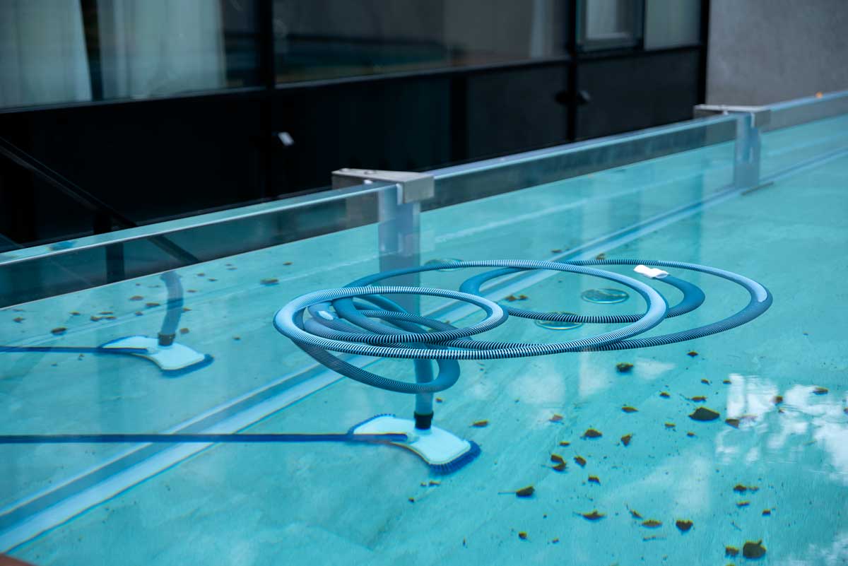 Ein Reinigungsbesen wird in das Wasser des Außenpools getaucht, um den Boden und die Wände zu reinigen; der Pool ist voller Blätter und weist andere Anzeichen von Vernachlässigung auf – Schmutz.