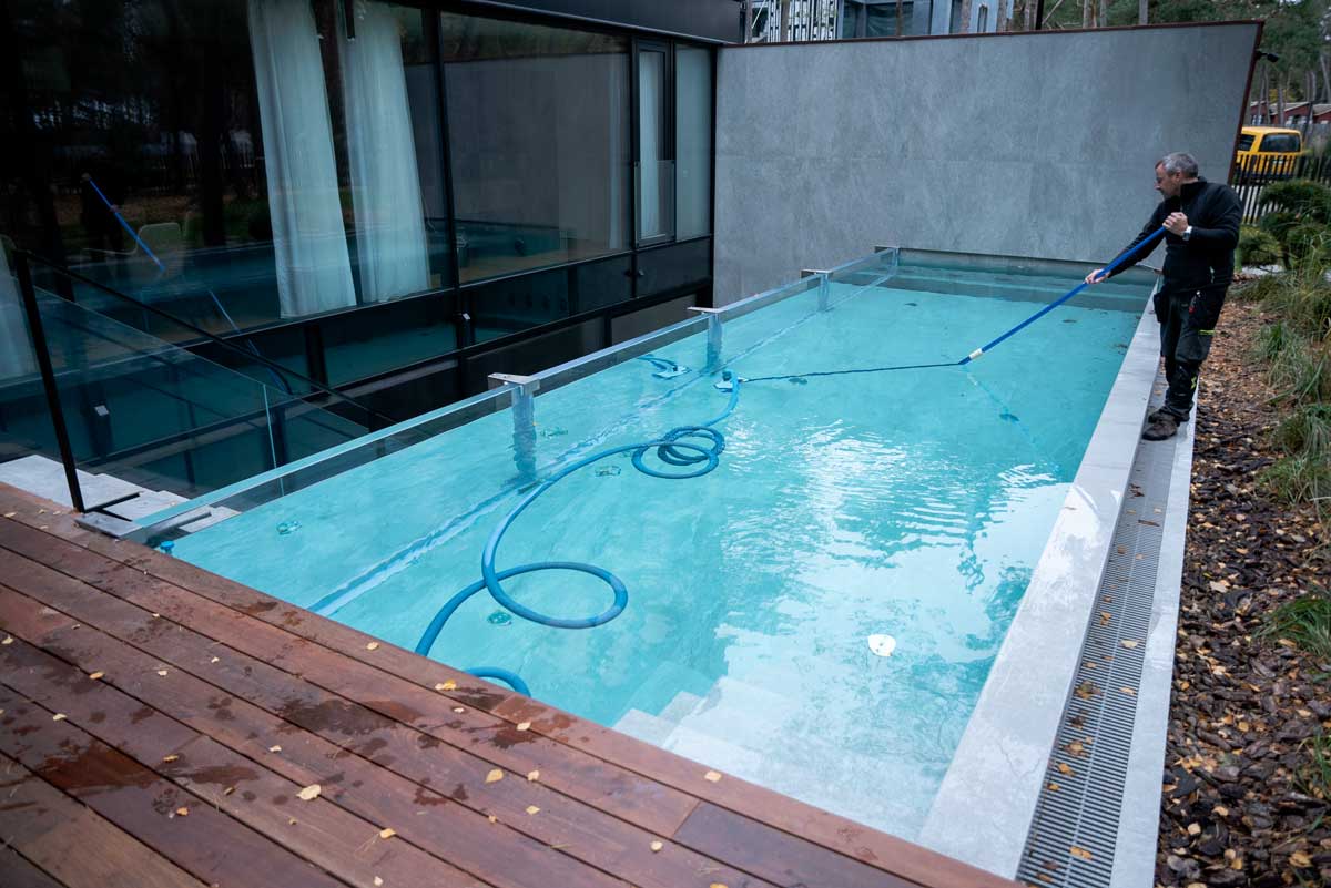Ein Schwimmbecken mit hellgrünem und trübem Wasser, dessen Oberfläche mit einer Algenschicht bedeckt ist; die Szene wurde eingefangen, während eine Person das Becken reinigt.