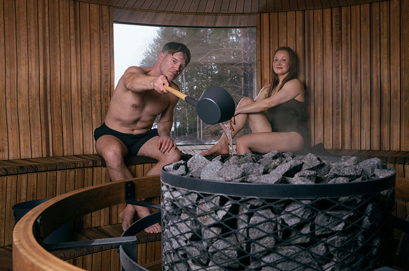 Ein Mann gießt mit einer Schöpfkelle Wasser auf die erhitzten Saunasteine, eine Frau sitzt daneben; eine dampfende, runde Holzsauna mit Fenster.